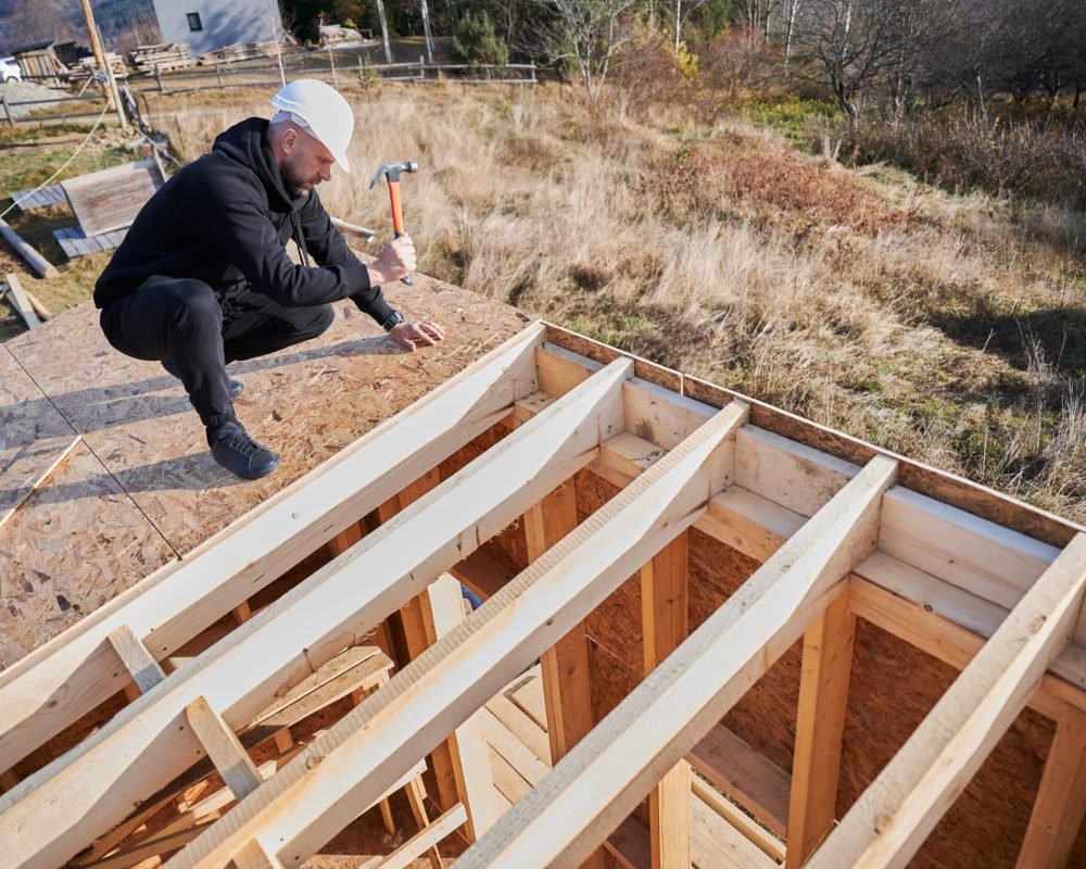 Carpenter hammering nail into OSB panel on the roof top of future cottage. Man worker building wooden frame house. Carpentry and construction concept.